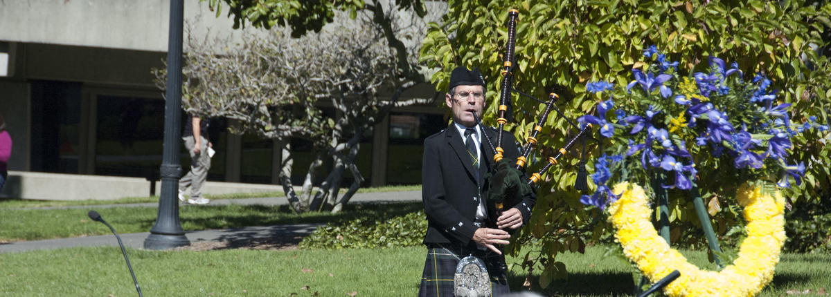 Photo of bagpipe player at campus memorial next to wreath of yellow and purple flowers
