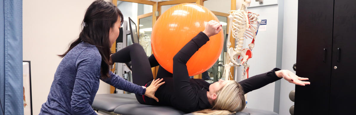 Student laying on back, holding yoga ball between legs and arm, one arm is stretched out. Physical therapist overseeing exercise.