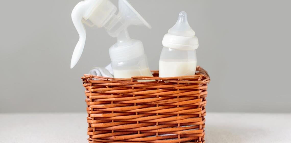 A brown wicker basket sits on a white table. The basket has a handheld breastfeeding pump and a baby bottle