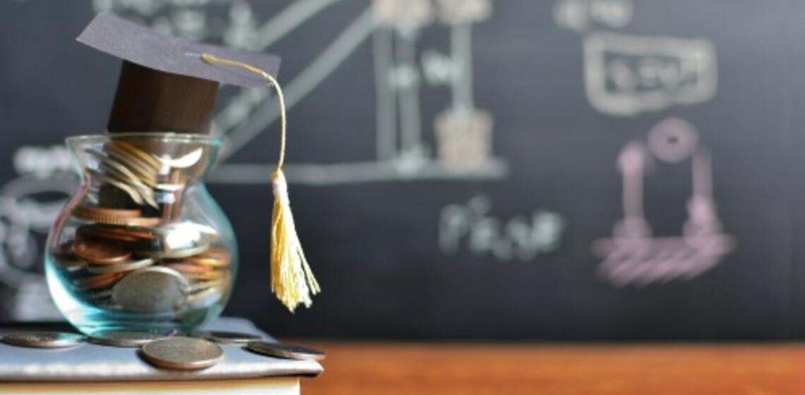 A money jar sits on a school desk. It has a graduation cap on it. There is a chalkboard with science equations behind it