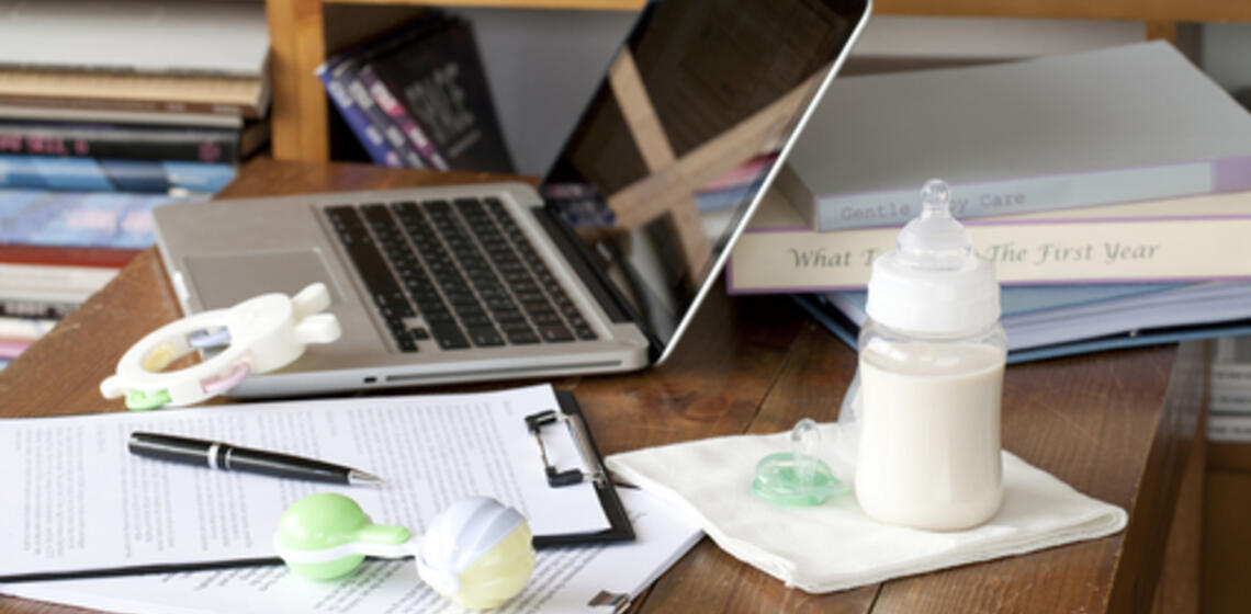 A wooden table top with a laptop, paper documents, and a baby bottle full of milk on it