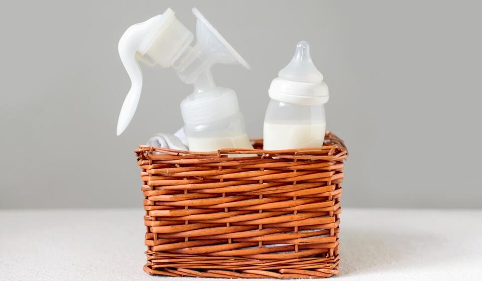 A basket sits on a white surface. It has a handheld breast pump and a baby bottle in it.