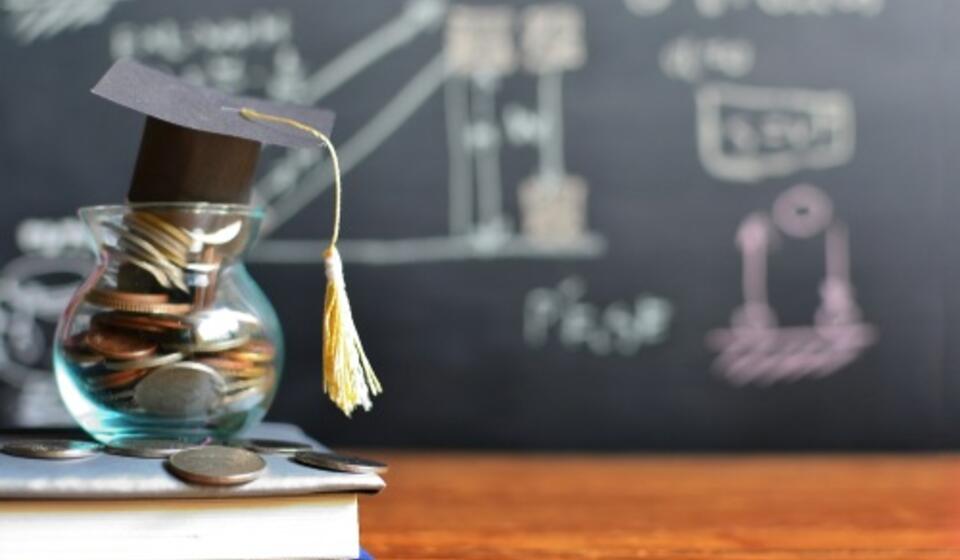 A text book sits on a desk with a jar of coins and a small graduation hat on top of it. A chalkboard with mathematics