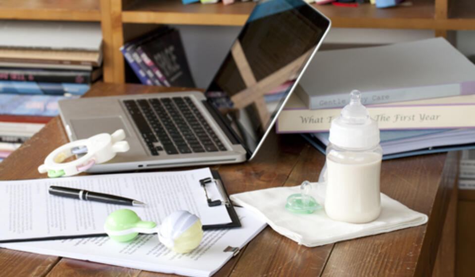 A table is full of papers, a baby bottle, rattle, and a laptop