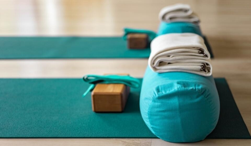 A yoga mat sits on a wooden floor. It has a yoga block, belt, and pillow on it